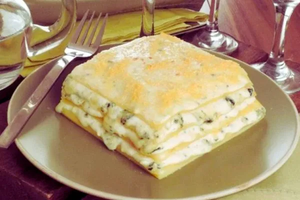 Lasanha de Queijos com Molho Gorgonzola Cremoso um pedaço de lasanha de queijos em cima de um prato com um garfo ao lado e taças de vinho