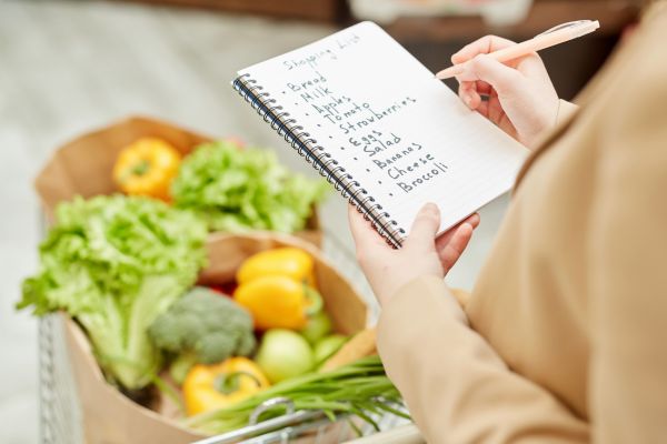 como fazer um cardapio semanal uma mulher com carrinho de compras com alimentos e um caderno na mão e um lápis com uma lista de alimentos