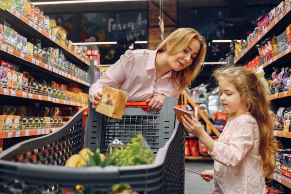 Como Fazer um Cardápio Semanal de Marmitas Congeladas para Ganhar Tempo e Comer Melhor mulher com a filha no supermercado escolhendo o que comprar com carrinho cheio de alimentos olhando para a filha que está olhando para o caderno